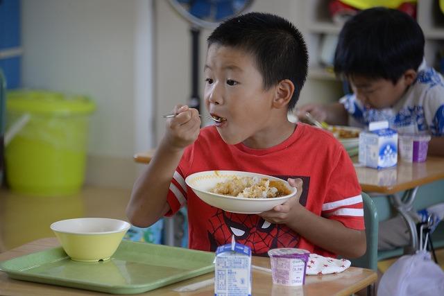 机に座って給食のカレーライスを頬張る赤いシャツを着た男の子の写真