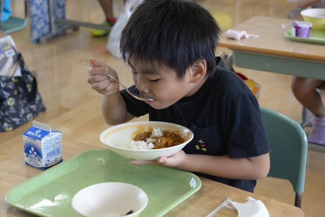 机に座って給食のカレーライスを頬張る黒いシャツを着た男の子の写真