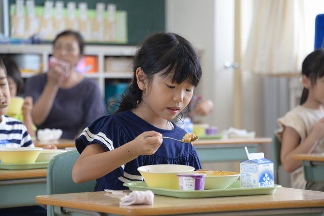 机に座って給食のカレーライスをスプーンですくって食べている青い服を着た女の子の写真