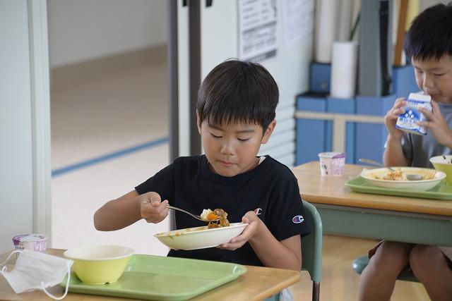 机に座って給食のカレーライスをスプーンですくって食べている黒いシャツを着た男の子の写真