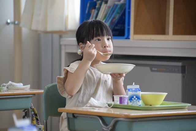 机に座って給食のカレーライスをスプーンですくって食べているベージュの服を着た女の子の写真
