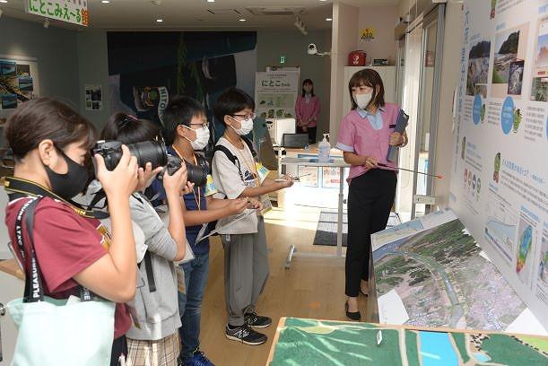 館内で解説をする女性職員とメモや撮影を行う人たちの写真