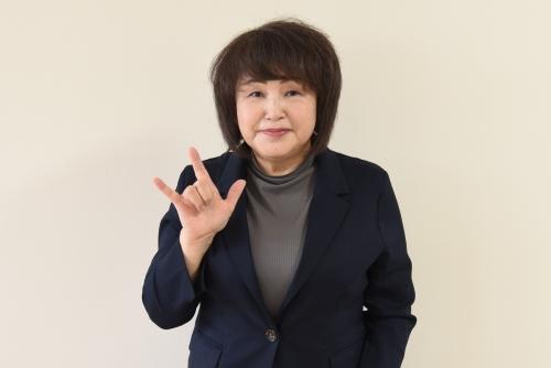 キツネのような手の形を見せる黒いスーツ姿の松田直子さんの写真