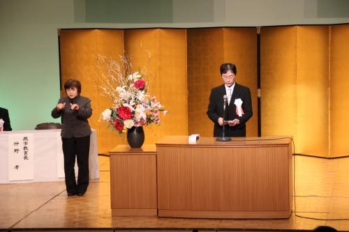 花が飾られた壇上の横で手話をする松田直子さんの写真