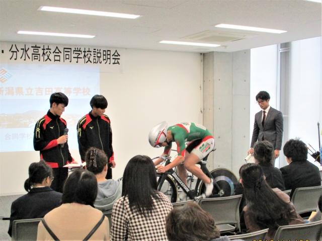 自転車をこぐパフォーマンスをする男性とそれを見守る人たちの写真