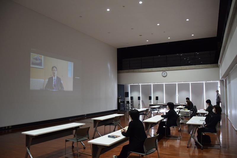 横長の机が並べられていて、その前に男性が話している姿が映されている大きなスクリーンがある写真